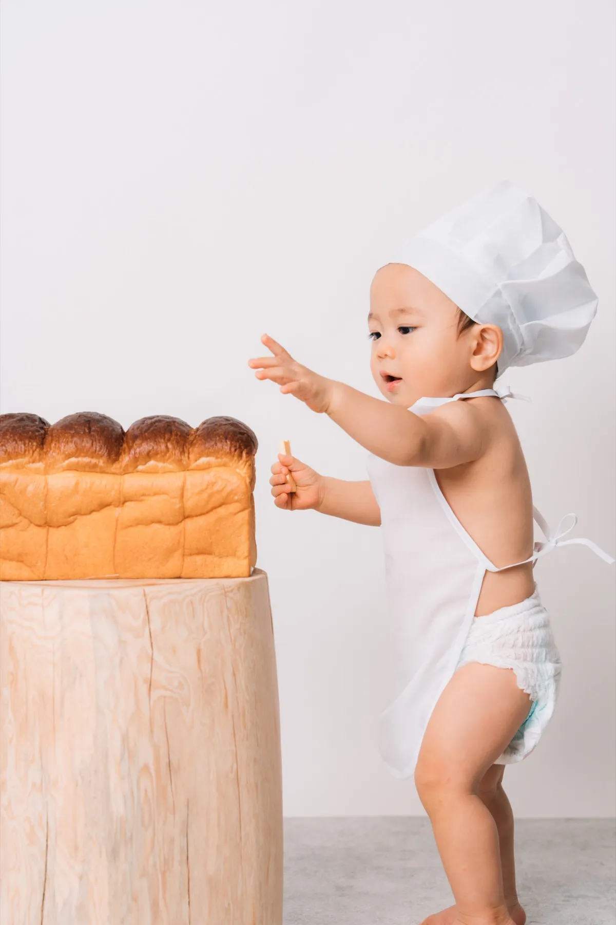 コック帽とエプロン姿で、大きな食パンに手をのばす1歳の赤ちゃん